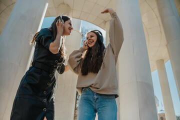 Low angle view shot of two brunette girls listening to different songs on their headphones, smiling and looking at each other. Having fun outdoors