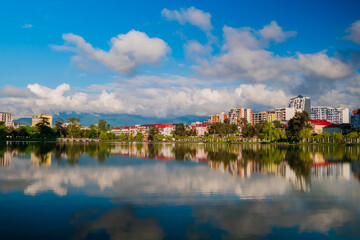 Fototapeta na wymiar View of the blue sky with white clouds over modern apartment, residential buildings with reflection in the water surface. 6 May Park on the shore of lake in Batumi in the afternoon