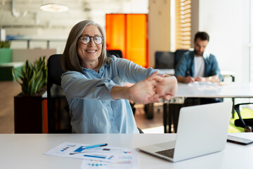 Portrait of smiling senior woman using laptop stretching arms, relax, taking a break working online in modern office. Successful confident businesswoman, entrepreneur finish work