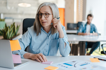 Smiling confident senior businesswoman using laptop, sticky notes looking at monitor in modern...