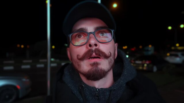 Portrait of man standing on the street and looking at billboard with flashing lights at night time. Closeup of male caucasian person with moustache staring at advertising reflecting in his eyeglasses.
