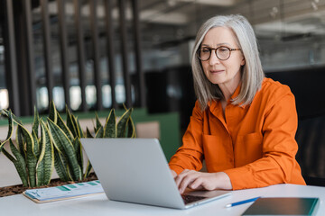 Confident serious mature businesswoman wearing stylish eyeglasses, orange shirt using laptop...