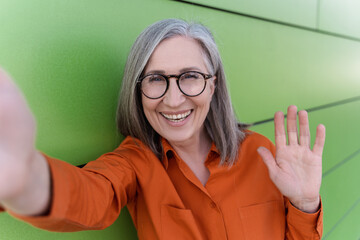 Happy grey haired senior woman taking selfie, waving hand, communication online, having video call standing outdoor. Smiling influencer recording video on the street. Technology concept