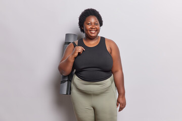 Body positive dark skinned woman dressed in black t shirt and leggings carries rolled rubber karemat going to have fitness training with coach poses against white background practices yoga every day