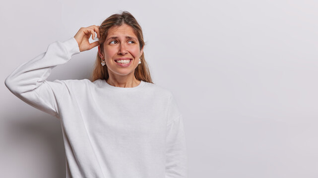 Waist up shot of confused European woman with long hair scratches head looks clueless clenches teeth dressed in casual sweatshirt poses against white background with copy space for your text