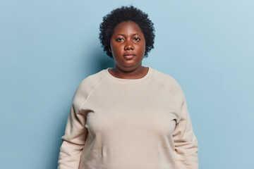 Portrait of serious dark skinned overweight young woman looks directly at camera dressed in sweatshirt concentrated at camera poses against blue background. Human face expressions and emotions