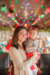 Fototapeta na wymiar Mother holding beautiful baby girl with colorful christmas lights in the background on train or bus