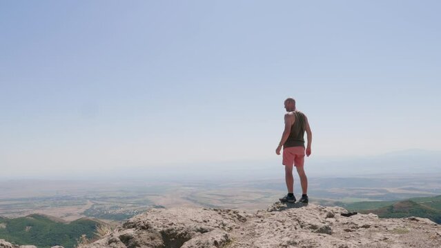 one bald guy walks along the edge on a high mountain on a bright sunny day, he goes from left to right, stops and looks around, goes further to the right