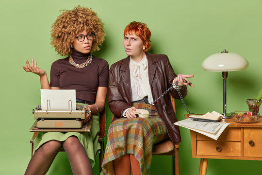 Back to past. Pair of young confused African american and European women sitting on chairs in old room typewriting wearing retro clothes on green background cant understand why phone isnt working