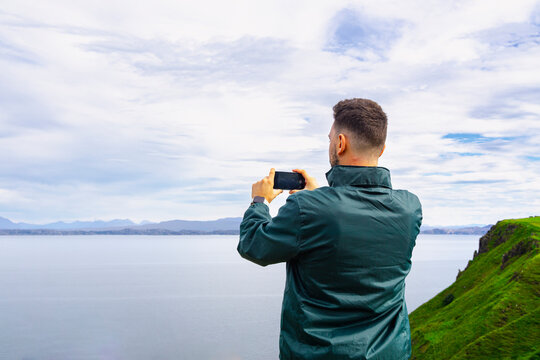 Young Caucasian man wearing a green jacket taking a photo with his smartphone of the Scottish Highlands landscape on the Isle of Skye.