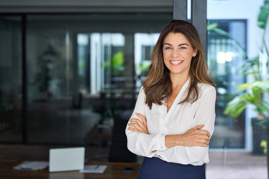 Happy cheerful 45 year old Latin professional mid aged business woman corporate leader, smiling positive mature female executive manager standing in office arms crossed looking at camera, portrait.