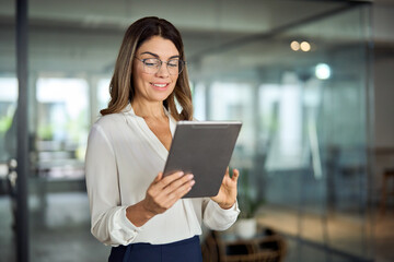 Smiling mid aged mature professional business woman banking manager, 40s female executive or entrepreneur holding fintech tab digital tablet standing in office at work, looking at pad. - obrazy, fototapety, plakaty