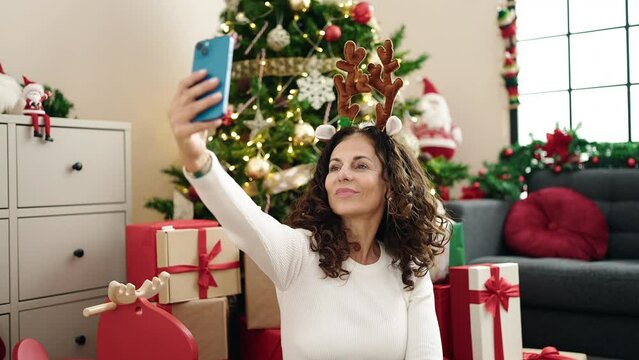 Middle age hispanic woman make selfie by smartphone sitting on floor by christmas tree at home