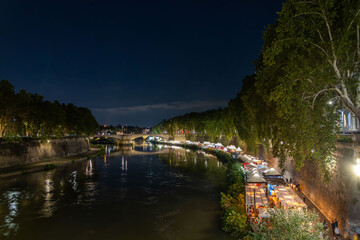 Fototapeta premium Vatican City in the distance with the Tiber river and pop-up restaurants on its banks, in Rome, Italy, at night