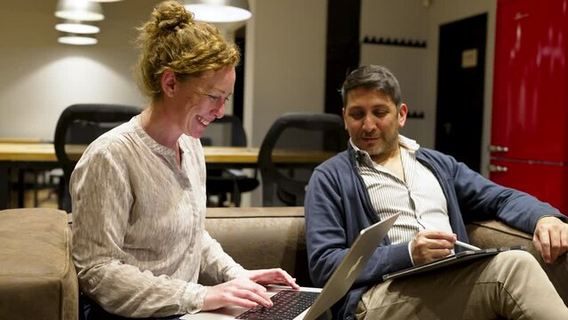 Two Business People In Coworking Space Interior Sitting On Sofa Having Conversation Using Laptop