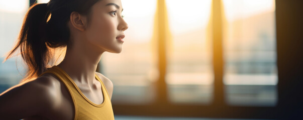 A Chinese woman dressed in a bright yellow athletic top and black yoga pants gazing out the window with a look of determination on her face her hands clasped together and a glowing