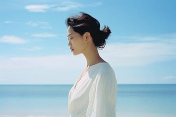 An Asian woman with a neat bun and white dress stands on an empty beach a clear blue sky stretching in the background. She gazes out with an air of serenity.