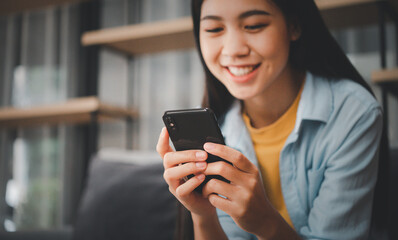 Smiling young asian woman using mobile phone checking social media,app playing game, shopping online, ordering delivery relax  while sitting on a couch at home with laptop computer