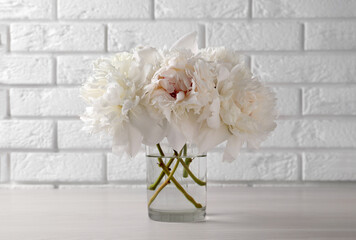 Beautiful peonies in glass vase on white table near brick wall