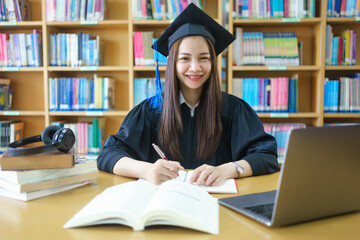 An asian girl student in university graduate gown with books, laptop studies happily in library.