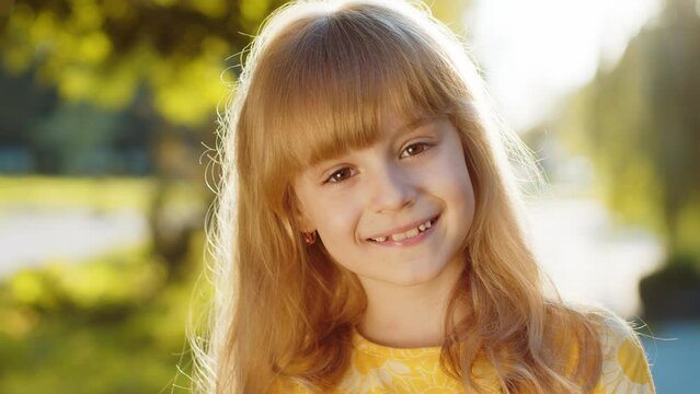 Portrait of happy preteen child kid smiling friendly face glad expression looking at camera winking with eye feel relaxation satisfied good mood. Pretty blonde girl on city sunset park street outdoors