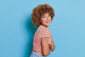 Side view of cute curly girl wearing pink top posing head to shoulder hugging herself with smile against blue background, wonderful time concept, copy space
