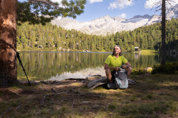 Adult gray-haired man with backpack sitting on the shore with his back to a mountain lake with a beautiful reflection