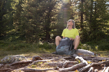 An adult man with a backpack sits in a sun-drenched clearing with tree roots