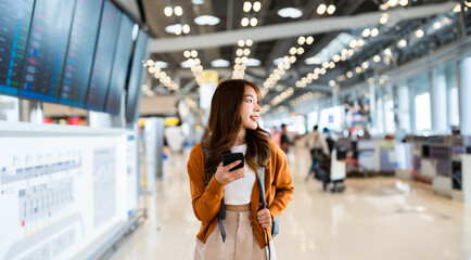Young asian woman in international airport, using mobile smartphone and checking flight at the flight information board - obrazy, fototapety, plakaty