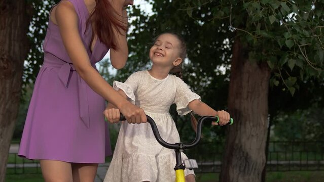 Smiling little daughter rides small bicycle with careful mother help in city park