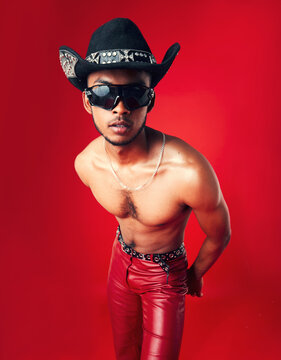 Fashion, Man And Cowboy In Hat, Shirtless With Sunglasses And Gen Z Isolated On Red Background. Confident Model, Cool And Leather Pants With Eyewear Accessory, Western Aesthetic And Edgy In Studio