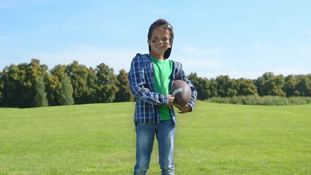 Portrait of cute African American school boy with eye black holding american football ball, looking with friendly cheerful smile, expressing positivity and happiness while playing game on green field.