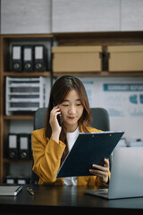 Portrait of Asian businesswoman talking mobile phone and working on laptop and papers on table in her office.