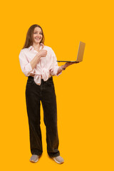 Young caucasian woman holds laptop computer in hand and shows thumbs up. Indoor studio shot, yellow background. Vertical frame. Copy space