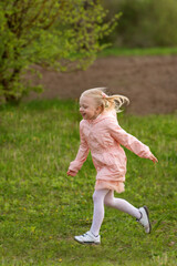 Portrait of little European girl with blonde hair in pink outfit running across field. Vertical frame.