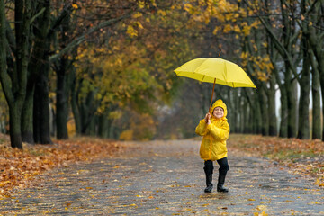 Happy cheerful boy in a yellow raincoat is jumping in the park with an umbrella. Autumn rainy day