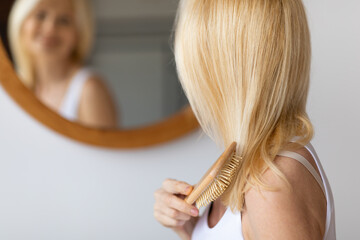 Rear view of middle aged woman brushing hair with wooden hairbrush looking at mirror in bathroom at...