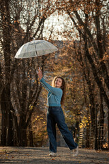 Student with wavy hair in autumn park holds transparent umbrella in hands. Girl looks directly into camera