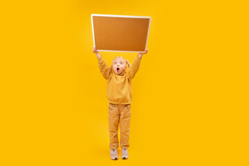 Little surprised girl in yellow suit holds blank board high above her head. Yellow background. Copy space.