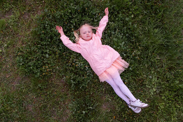 Little smiling girl in a pink dress lies on a green lawn with her arms outstretched and resting. Spring holidays