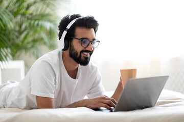 Young Indian Guy Wearing Headphones Resting In Bed With Laptop And Coffee