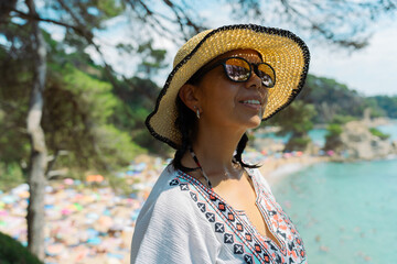 Attractive latina woman with sunglasses and hat enjoying from the top of the mountains some beautiful views of a beach on the Costa Brava with bather on a beautiful summer day