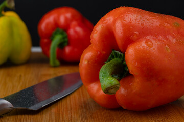 Three peppers with a knife on a wooden chopping board. Bell peppers of various colors on a wooden cutting board on a black background.