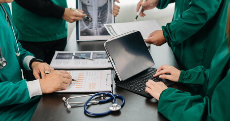 Medical team having a meeting with doctors in white lab coats and surgical scrubs seated at a table discussing a patients working online using computers in the medical