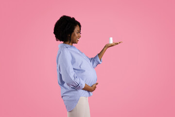 Cheerful young black pregnant woman touches big belly, enjoys motherhood, show jar of pills, vitamins and minerals
