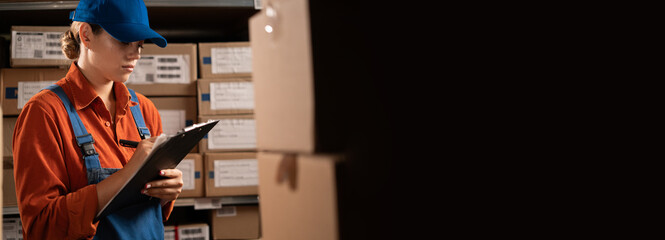 Female warehouse worker working with clipboard writing down inventory numbers of parcel boxes
