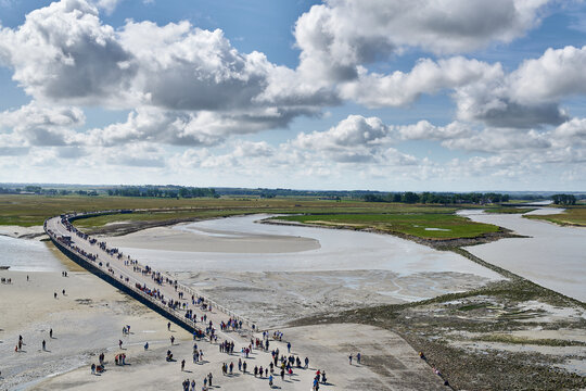 Wooden walkway full of people crossing a bay without water with a green forest in the background, water and the sky covered with white clouds