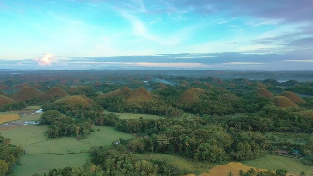 Chocolate Hills at sunset in Bohol Island, Philippines. Aerial drone cinematic shot. Backward travelling above landmark and nearby Carmen village. Vibrant dusk colors and dramatic cloudy sunset.