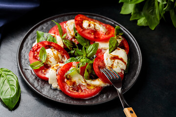 Caprese Salad. Tomato and mozzarella slices with basil leaves with fork on dark plate. Traditional Italian food