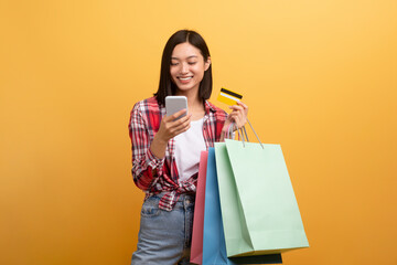 Cool smiling asian lady in casual holding colorful shopping bags, smartphone and bank card, yellow studio background
